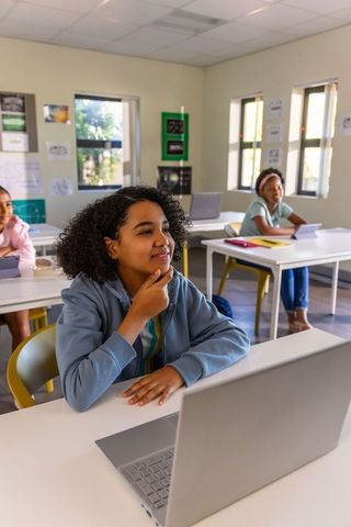 Thinking preteen girl using laptop in modern classroom with diverse classmates learning