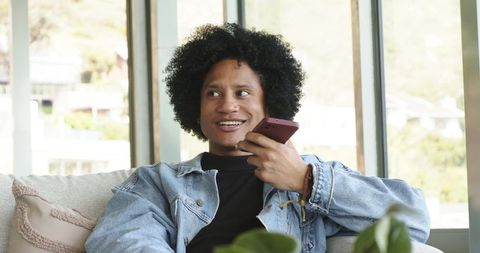Smiling african american man in denim jacket holding smartphone on living room sofa
