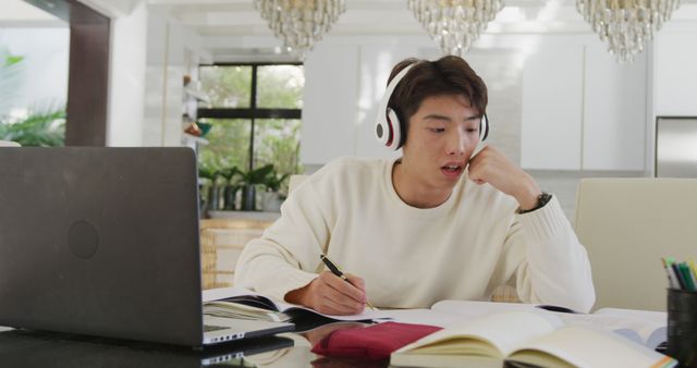 Asian male teenager with headphones learning and using laptop in living room. spending time alone at home.