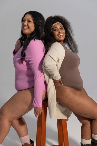 Diverse women sitting back-to-back wearing bodysuits and cardigan, smiling confidently