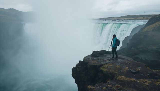 Hiker capturing waterfall from cliff edge with tripod in misty rugged gorge