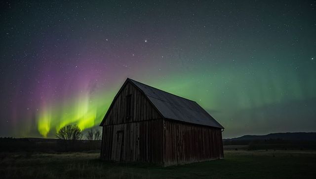 Rustic weathered barn glowing beneath northern lights over pasture and starry sky
