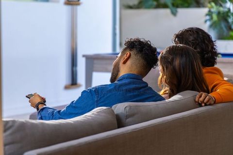 Diverse friends relaxing on couch watching tv, holding remote, cozy daytime living room