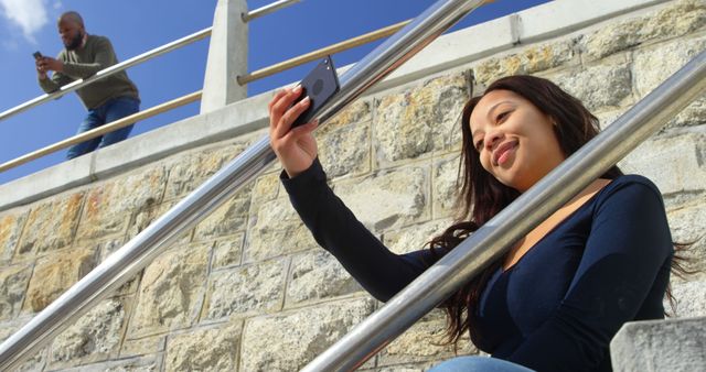 Woman taking selfie on metal stairs while man using phone in the background. Great for illustrating social media, technology use, modern lifestyle, outdoor activities or casual moments.