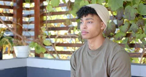 Young african american man sitting on patio bench wearing beanie and olive sweatshirt, contemplative