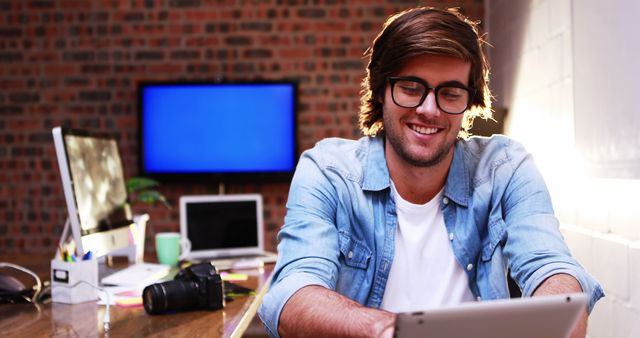Young man is using tablet and smiling in a trendy office with exposed brick walls. He is wearing glasses and casual clothing, suggesting a modern creative workspace. There are a laptop and camera on his desk. Perfect for illustrating themes of technology, modern workspaces, creative industries, or entrepreneurship.