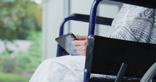 Person sits in wheelchair while holding mobile device. Photo captures moment of independence and connectivity. Can be used for topics of disability, technology integration in daily life, and outdoor leisure for people with mobility challenges.