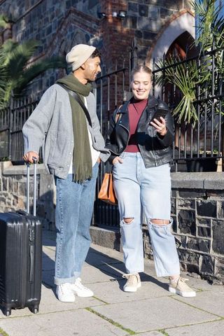 Young couple walking historic city street pulling suitcase and checking smartphone