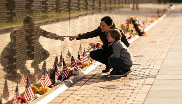 Mother and son pointing at engraved names on veterans memorial wall with flags