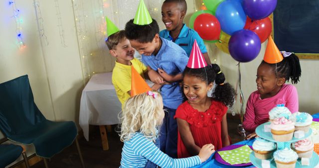 A diverse group of children celebrates a birthday party, with colorful balloons and party hats enhancing the festive mood. Their joyful expressions and the cake on the table suggest a moment of happiness and celebration.