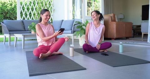Diverse women friends laughing on patio yoga mats using smartphones post-workout