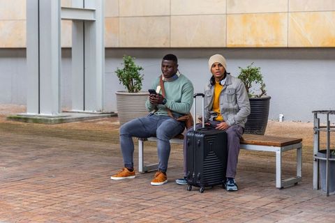 Diverse male friends sitting on bench checking phone next to suitcase in urban plaza