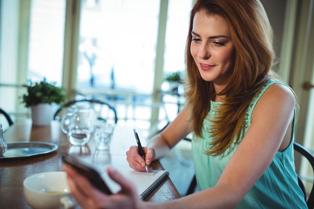 Woman using mobile phone while writing on notepad in cafÃ©