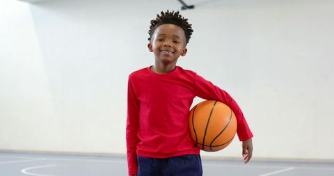Smiling african american boy holding basketball on indoor court wearing red shirt confident