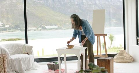 Mature woman mixing paint in bright coastal studio with ocean view and canvas on easel