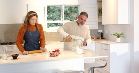 Couple baking together in modern white kitchen, man pouring wine while woman rolling dough