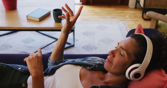 Woman wearing headphones, relaxing on couch, enjoying music. Ideal for lifestyle, leisure, or relaxation themes. Can be used in blog posts, website banners, advertisements promoting home comfort, music streaming services, or mental wellness tips.