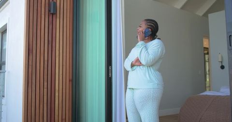 African american woman in mint pajamas talking on phone at bedroom balcony doorway