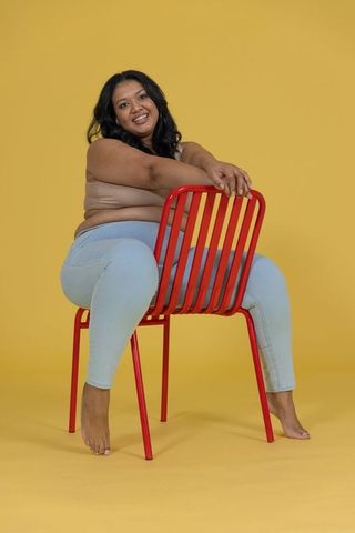 Curvy woman sitting backward on red chair smiling on vibrant yellow backdrop, body positive denim