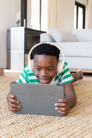 African american boy lying on woven rug using tablet with headphones for learning and play