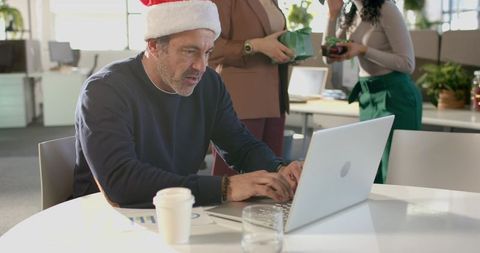 Mature man wearing Santa hat typing on laptop during festive office party with coworkers