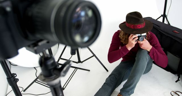 Photographer wearing hat sitting on floor of studio with camera tripod in foreground. Professional photography equipment visible, suggesting an indoor photo shoot. Ideal for use in articles and websites about professional photography, studio setups, or creative professions.