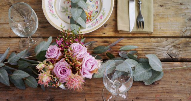 Rustic table setting with pink roses in a centerpiece, floral dishware, glassware, cutlery, and eucalyptus leaves on a wooden table. Ideal for use in articles or promotional materials related to dining experiences, wedding planning, rustic decor, or floral arrangements.