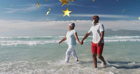 Active senior couple walking hand in hand through surf under floating gold stars