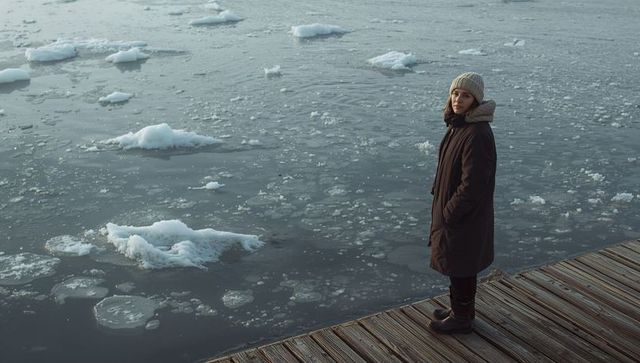 Solitary woman standing on icy dock wearing winter coat and knit beanie by frozen harbor