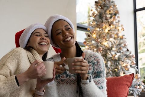 Friends wearing santa hats sharing hot drinks on couch by festive christmas tree