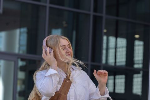 Blonde woman listening to music with pink headphones and brown leather bag by glass building