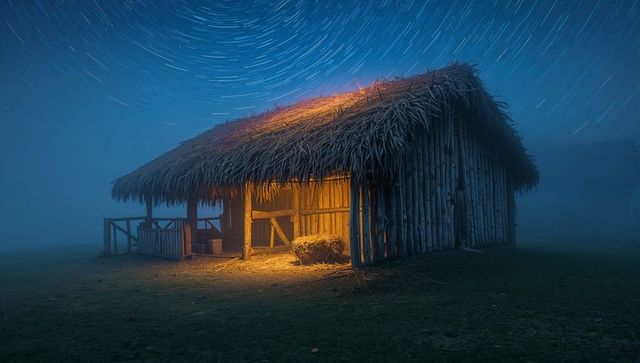 Glowing thatched hut with star trails on foggy night lantern-lit rustic cabin hay bale