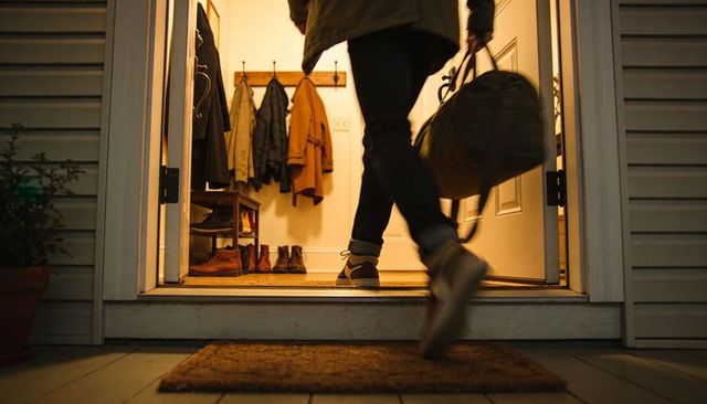 Traveler entering home at dusk carrying duffel bag crossing front door threshold into warm foyer