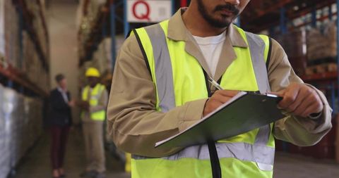 Warehouse worker writing on clipboard wearing hi-vis vest checking inventory on pallet racking