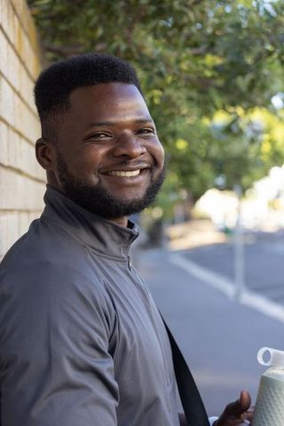 Smiling urban man leaning on brick wall holding water bottle and backpack strap