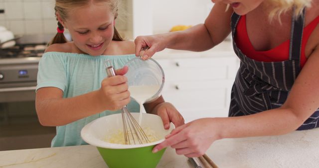 Mother and daughter engaged in cooking activity in a modern kitchen. Daughter is smiling while whisking ingredients in a green bowl, with her mother gently guiding her. This warm and joyful scene can be used for advertisements promoting family activities, cooking products, and educational content on cooking with children.