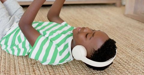 African american boy listening with headphones lying on jute rug using tablet