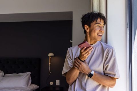 Asian man holding smartphone by sunlit bedroom window smiling in modern minimal interior