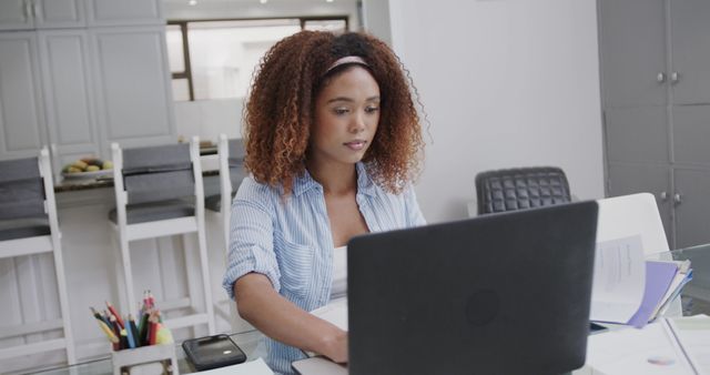 Young woman with curly hair working remotely from home using laptop in modern kitchen. Suitable for use in articles about remote work, studying, productivity tips, home office setups, and freelance careers.