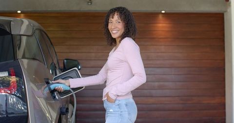 Smiling Woman Charging Electric Car in Garage with Wooden Background