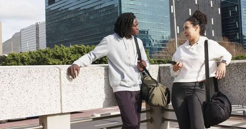Diverse friends leaning on urban railing chatting while carrying duffel bags and gym gear