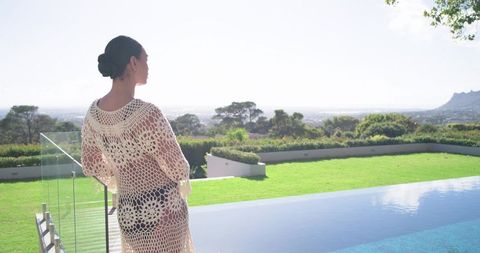 Woman standing on infinity pool rim wearing crochet coverup facing scenic hills and horizon