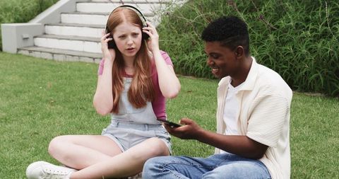 Teen Friends Sharing Headphones and Smartphone on Campus Lawn, Listening and Laughing