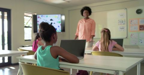 African girl using laptop in modern classroom while teacher presenting interactive lesson