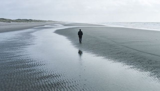 Solitary man walking along misty windswept beach leaving footprints by tidal channel