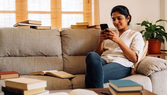 Young woman relaxing on sofa using smartphone surrounded by books and cozy home decor