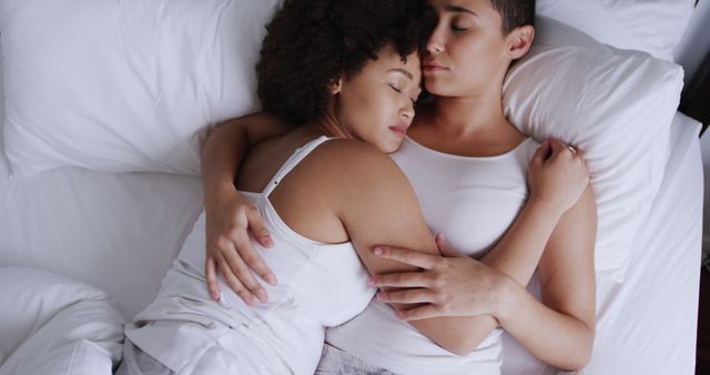 Interracial lesbian couple embracing lovingly in bed, wearing white shirts. Represents unity, love, and comfort in a diverse relationship. Ideal for themes about love, affection, LGBT rights, diversity, and intimacy in relationships.