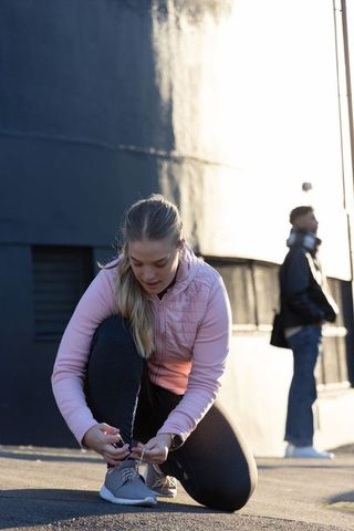 Teen female tying running shoe on urban plaza at sunrise wearing pink jacket and leggings