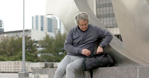 Mature man wearing headphones sitting on ledge rummaging in duffel bag by metal sculpture