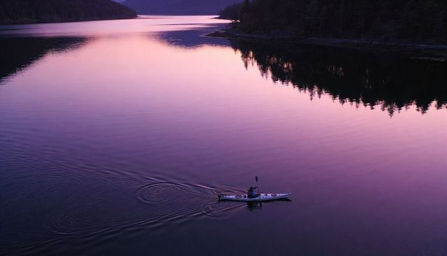 Solo kayaker paddling across glassy purple lake at dusk with forest reflection
